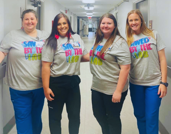 4 Lab workers standing in the hallway of the hospital. Wearing &quot;Saved by the Lab&quot; shirts and scrub pants.